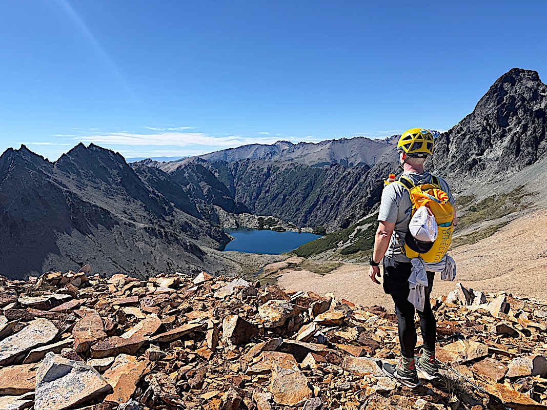 Helmeted hiker on loose scree above a deep blue mountain lake