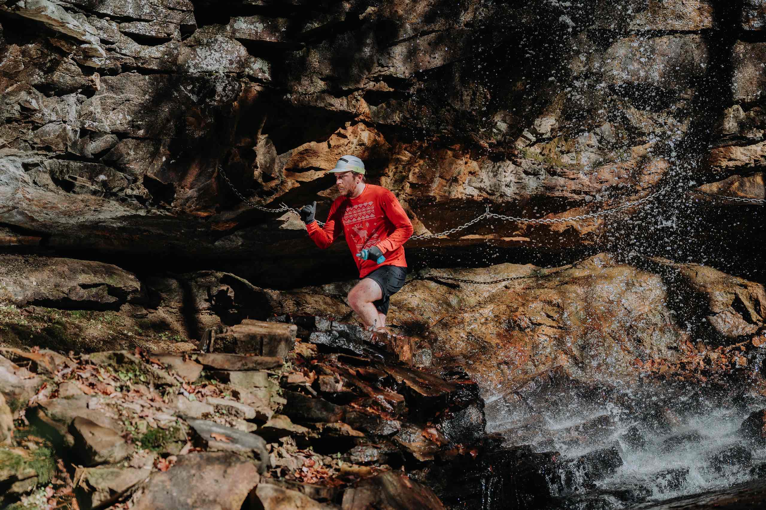A runner in a red shirt crosses wet rock beneath a waterfall on the Fiery Gizzard Trail