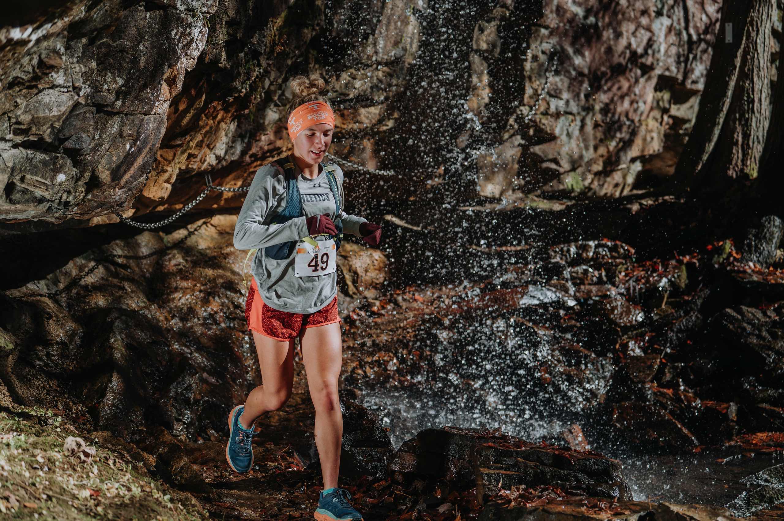 A runner moves through bright waterfall spray against layered rock