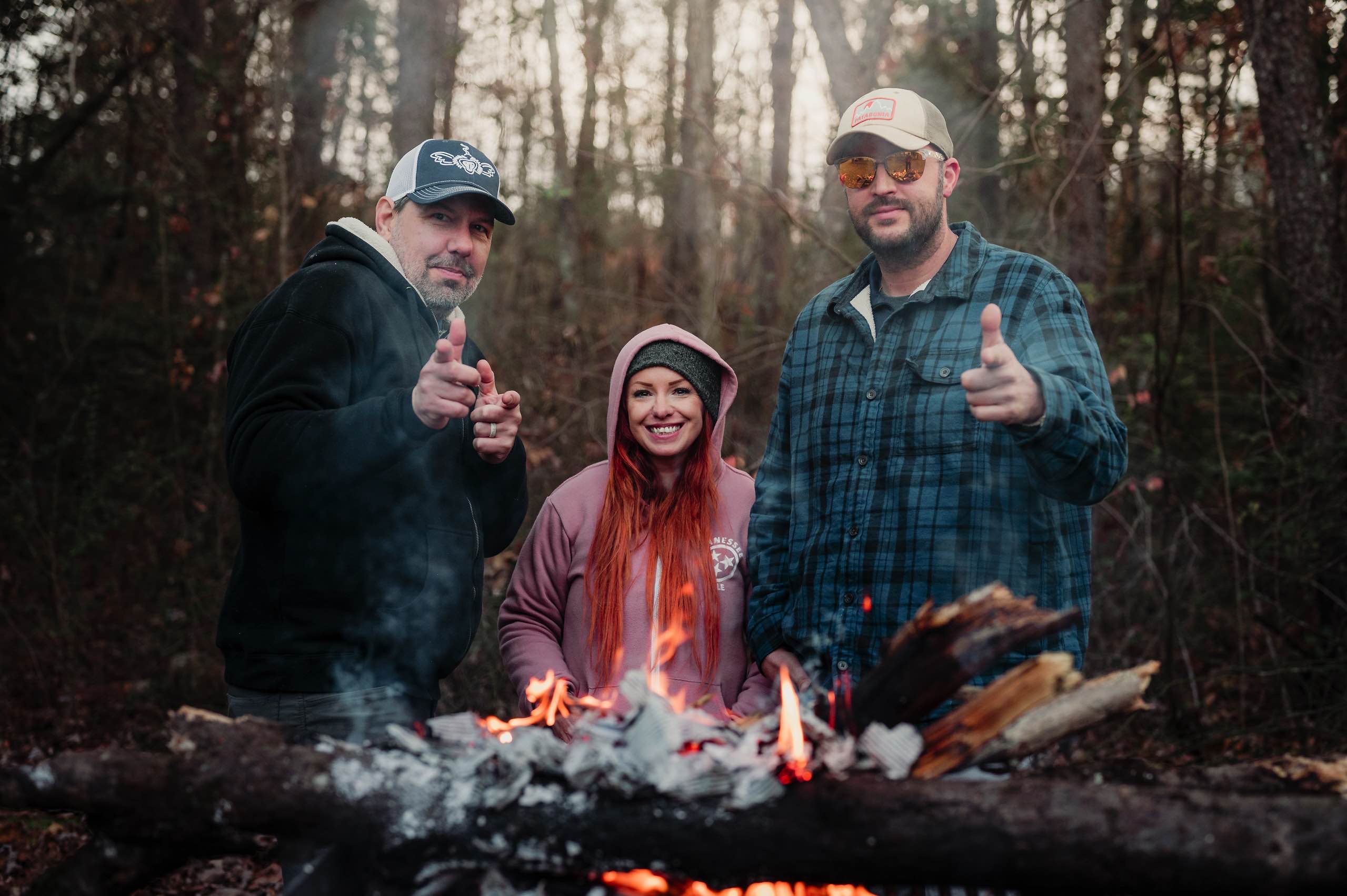 Three Fiery Gizzard volunteers smile behind a campfire in the winter woods