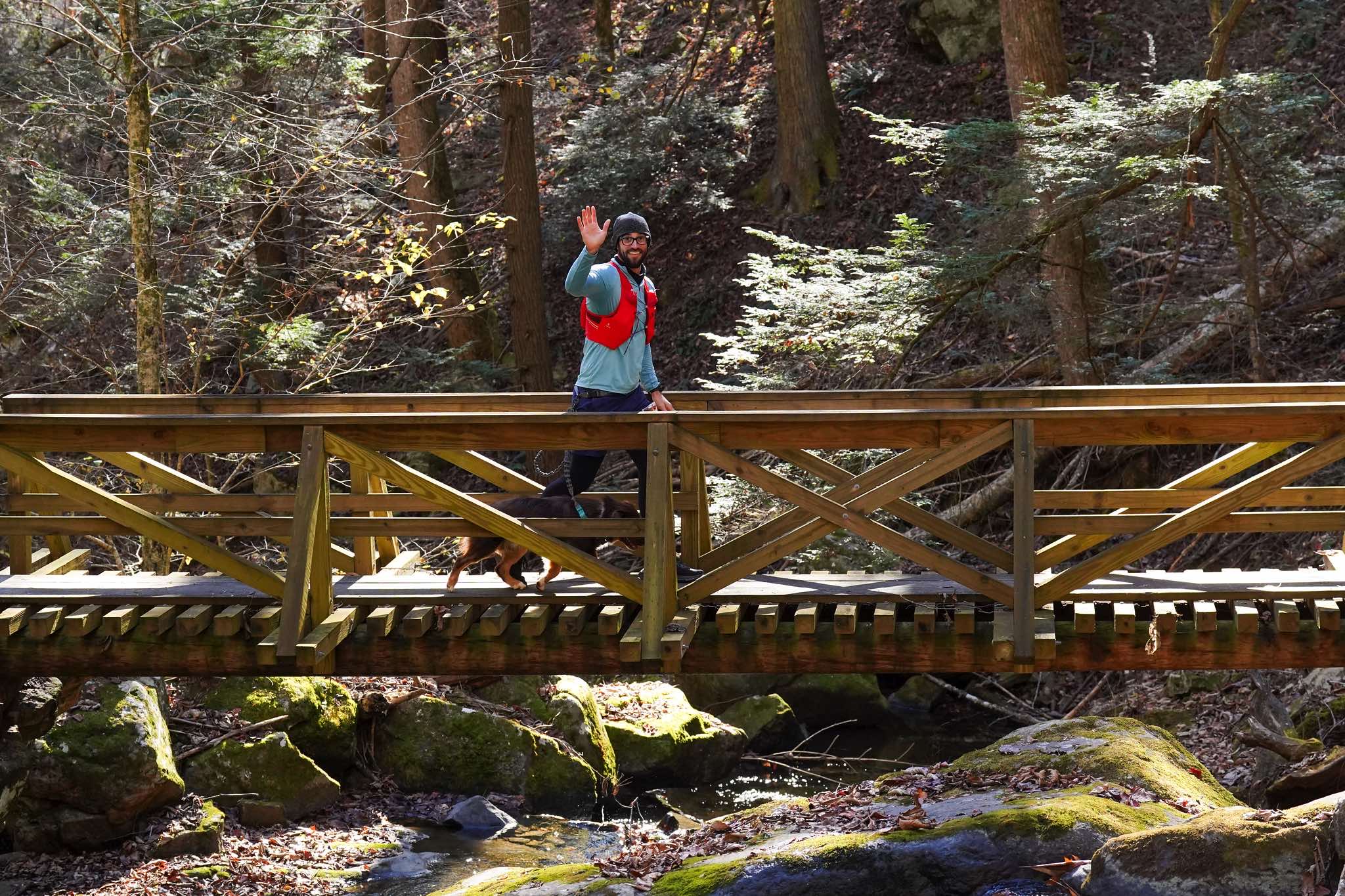 A runner waves while crossing a wooden bridge with a dog