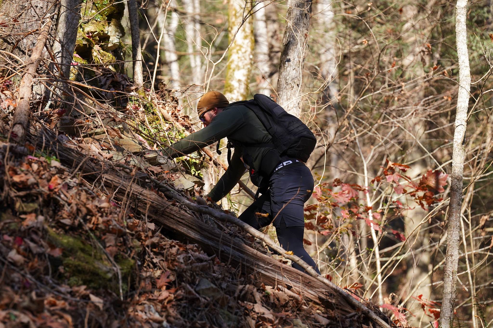 A runner uses both hands to climb a steep leaf covered slope