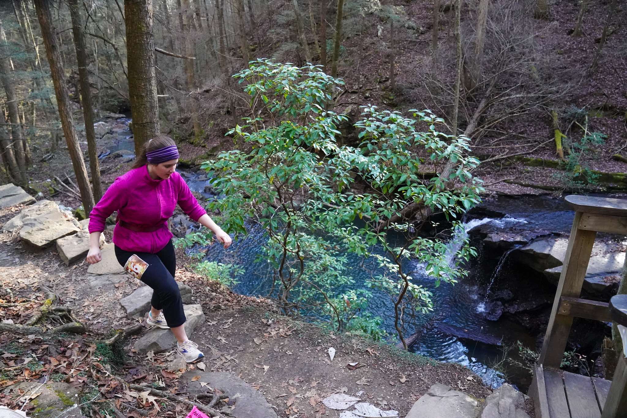 A runner climbs trail steps above a creek and cascade